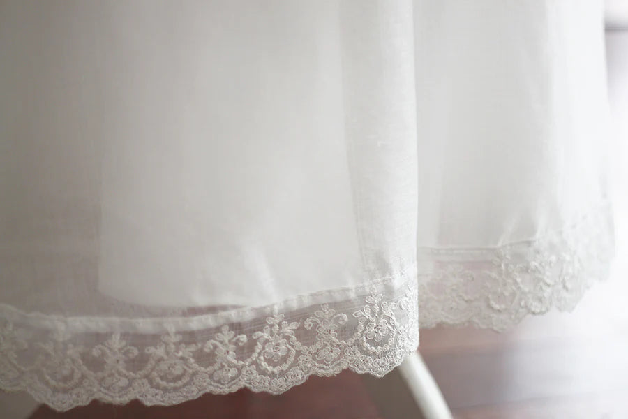 Cape de baptême longue traditionnelle pour baptême en mousseline blanc cassé et dentelle fine. Se ferme avec un large ruban. Fait main en France. Gros plan sur le bas de la cape.