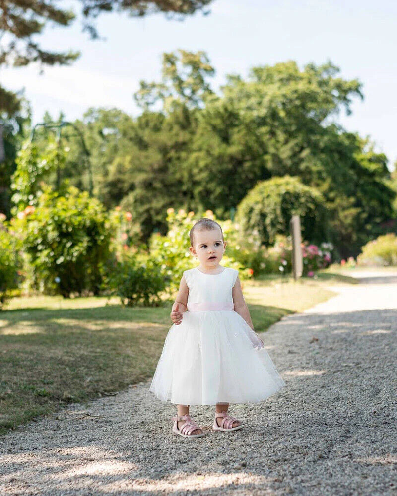 Robe de baptême ou de cérémonie pour bébé fille en coton blanc cassé avec une jupe en tulle et une ceinture rose poudré. Photo de la robe portée par une petite fille.