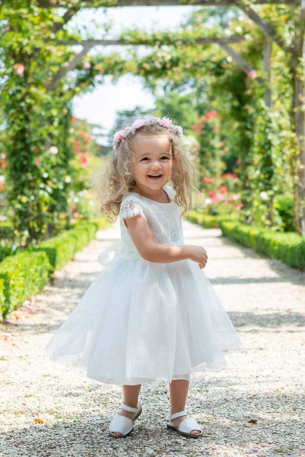 Robe de baptême et de cérémonie pour bébé fille, corsage en dentelle italienne blanc cassé, jupe en tulle pailleté, ceinture en soie. Robe de baptême haut de gamme, création française Fil de Légende. Photo de la robe portée par une petite fille qui marche.